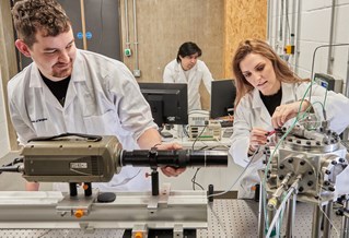 Three researchers wearing white lab coats, working with machinery and digital screens in the 㽶ֱ Advanced Engineering Centre